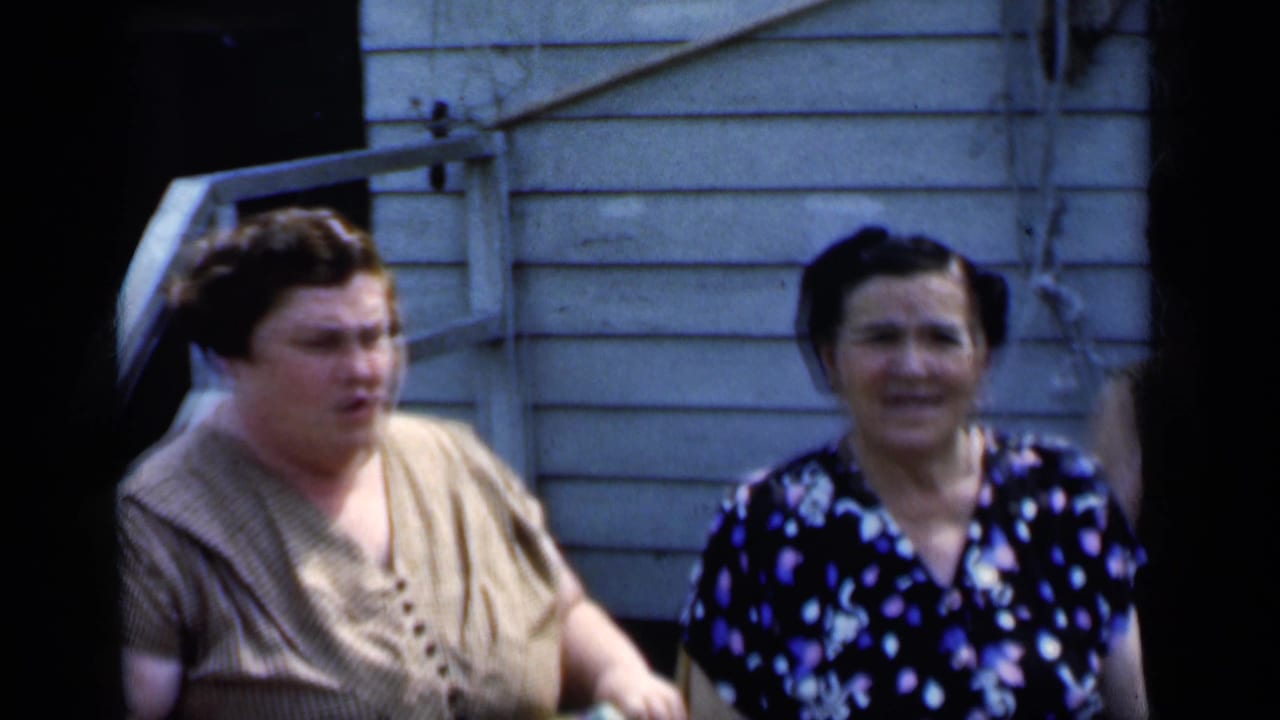 Two adult women stand outside a light clapboard building, one in beige and one in a dark floral dress.