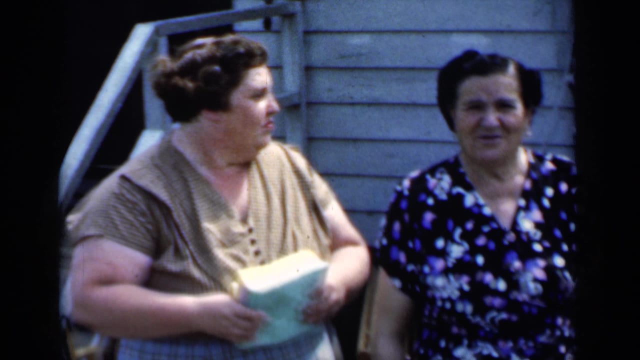 In the stated New York home movie, two women stand outside as one holds a light-colored book or papers.