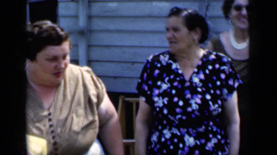Three adult women are visible outside the clapboard building, with one background woman wearing sunglasses.