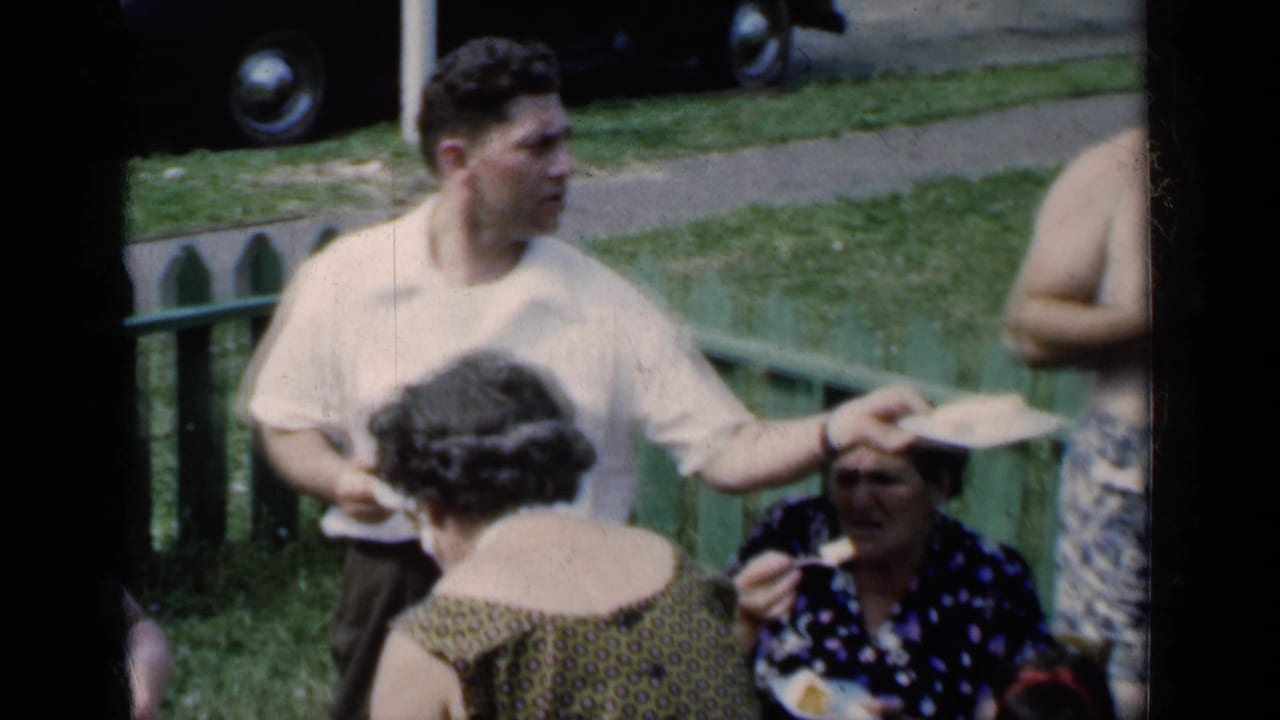 1951 New York outdoor gathering with a standing man holding a plate, women nearby, a green fence, grass, and part of a car in the background.