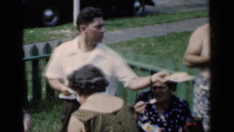 1951 New York outdoor gathering with a standing man holding a plate, women nearby, a green fence, grass, and part of a car in the background.