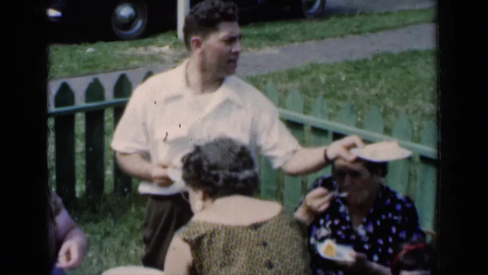 Candid 1951 New York home-movie view of adults sharing food near a green picket fence, with a seated woman eating and a car partly visible behind them.