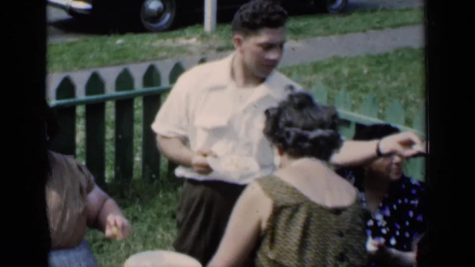 1951 New York summer-party frame with a man holding food, women nearby, a green fence, grass, and soft 8mm color and grain.