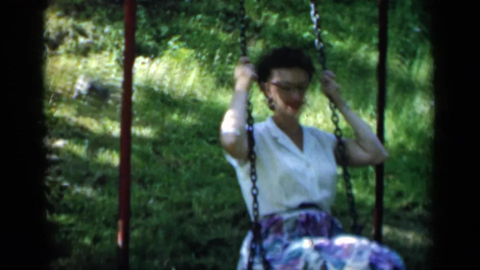 In 1951 New York home-movie footage, a woman with glasses holds a chain swing while wearing a white blouse and floral skirt.