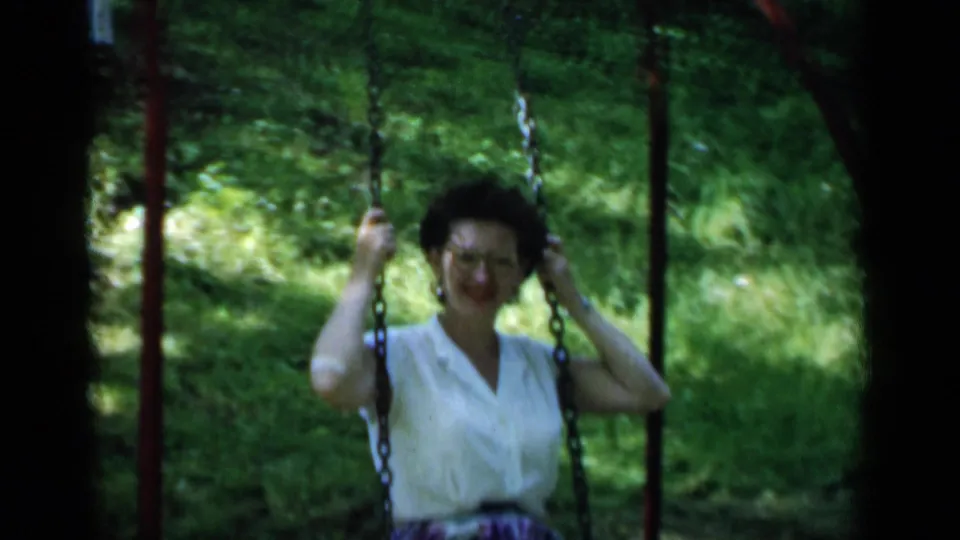 The woman smiles toward the camera while holding the chain swing, with dense greenery behind her in the 1951 New York footage.