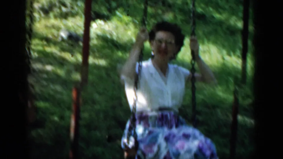 The woman swings forward in a white blouse and floral skirt, softened by 8mm motion blur and dappled outdoor light.