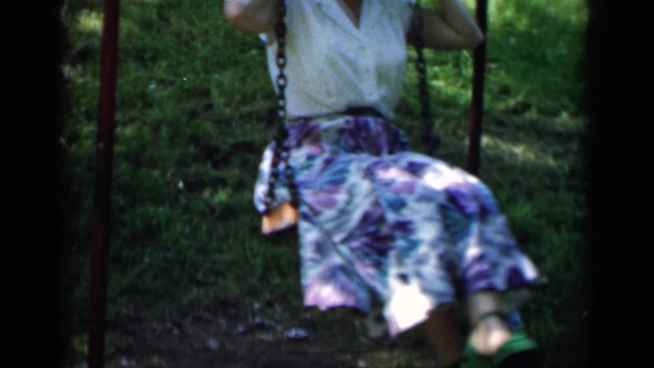Woman in a white blouse and floral skirt sits on a swing in a shaded grassy area.
