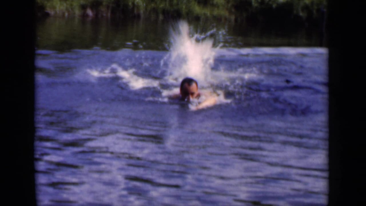 Adult man swimming through blue water with a splash behind him and green shoreline above.
