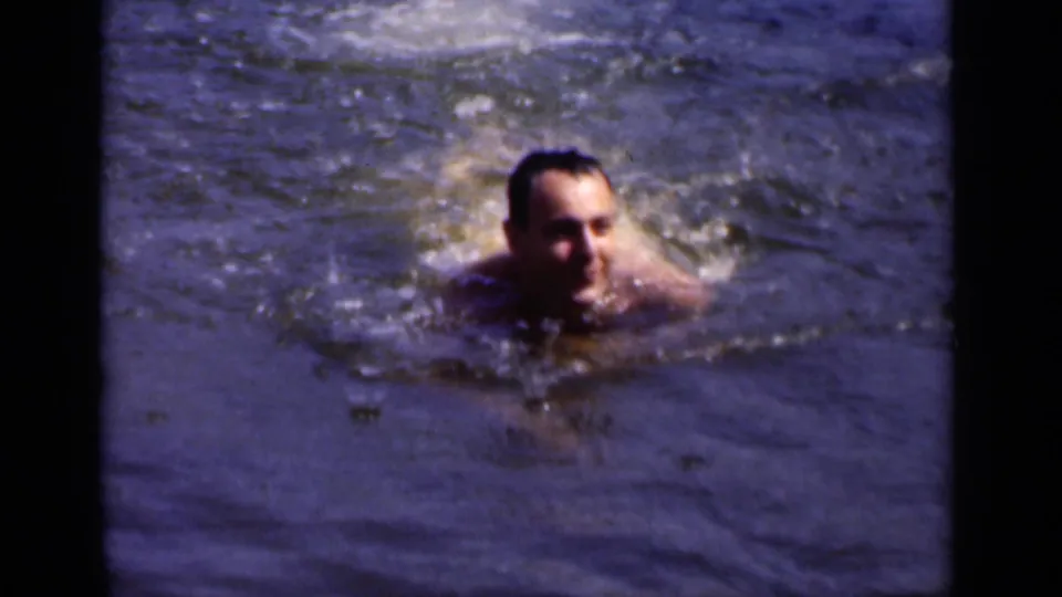 In 1951 New York home-movie footage, an adult man swims toward the camera with his face above rippling water.