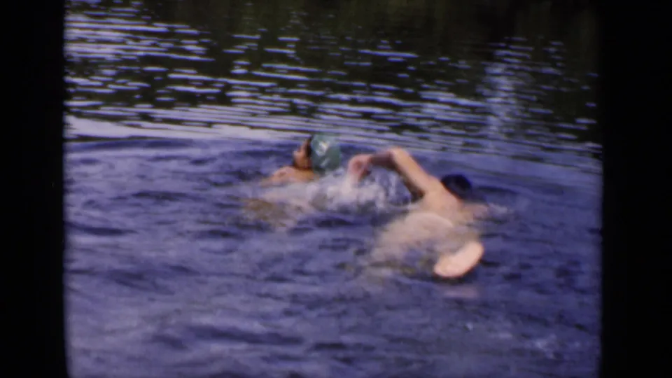 Two adults swim side by side in open water, with one swimmer wearing a green cap in this 1951 New York home-movie frame.