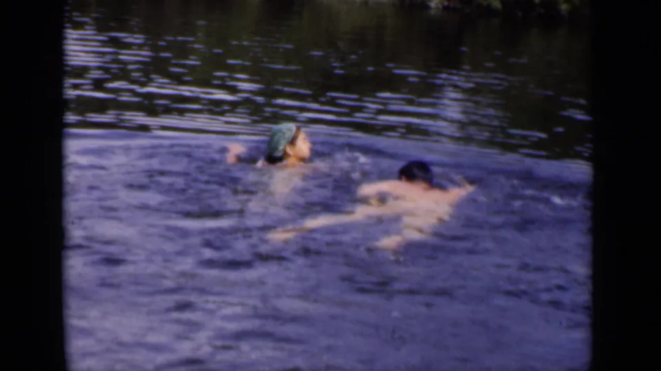 Two adults continue swimming across rippling water in 1951 New York footage, one wearing a green swim cap.