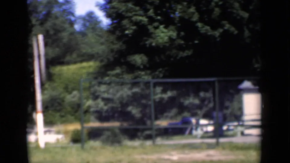 1951 New York exterior view with chain-link fencing, trees, a blue car, grass, and a light structure beside the property.