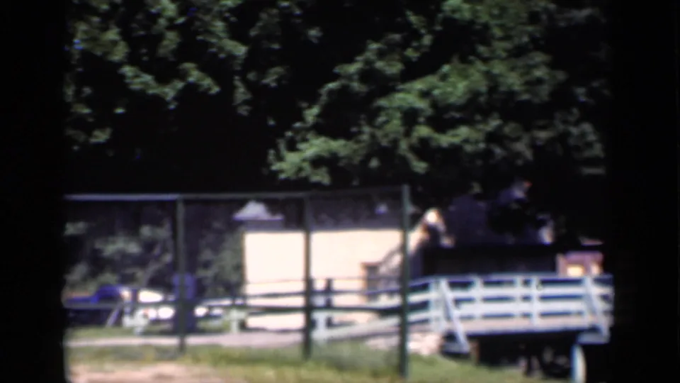 1951 New York home-movie frame showing a fenced hotel property with a parked blue car and small white bridge under trees.