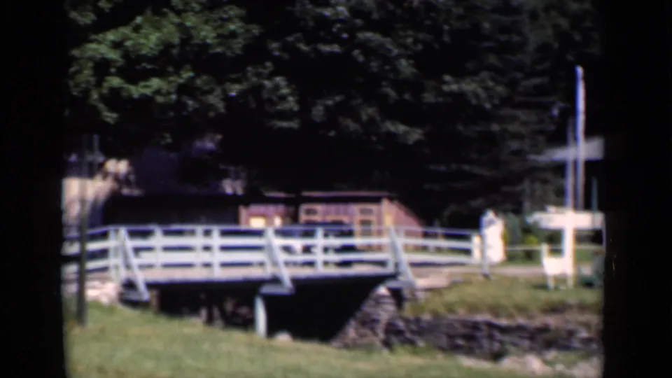 1951 New York exterior showing a small white bridge over a creek or river, with trees, grass, posts, and a person at right.