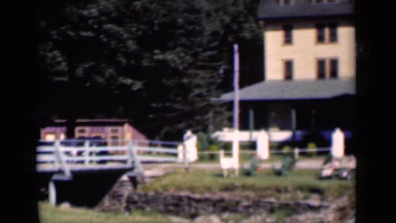 Yellow-orange building behind a white fence and stone wall, surrounded by trees in bright sun.