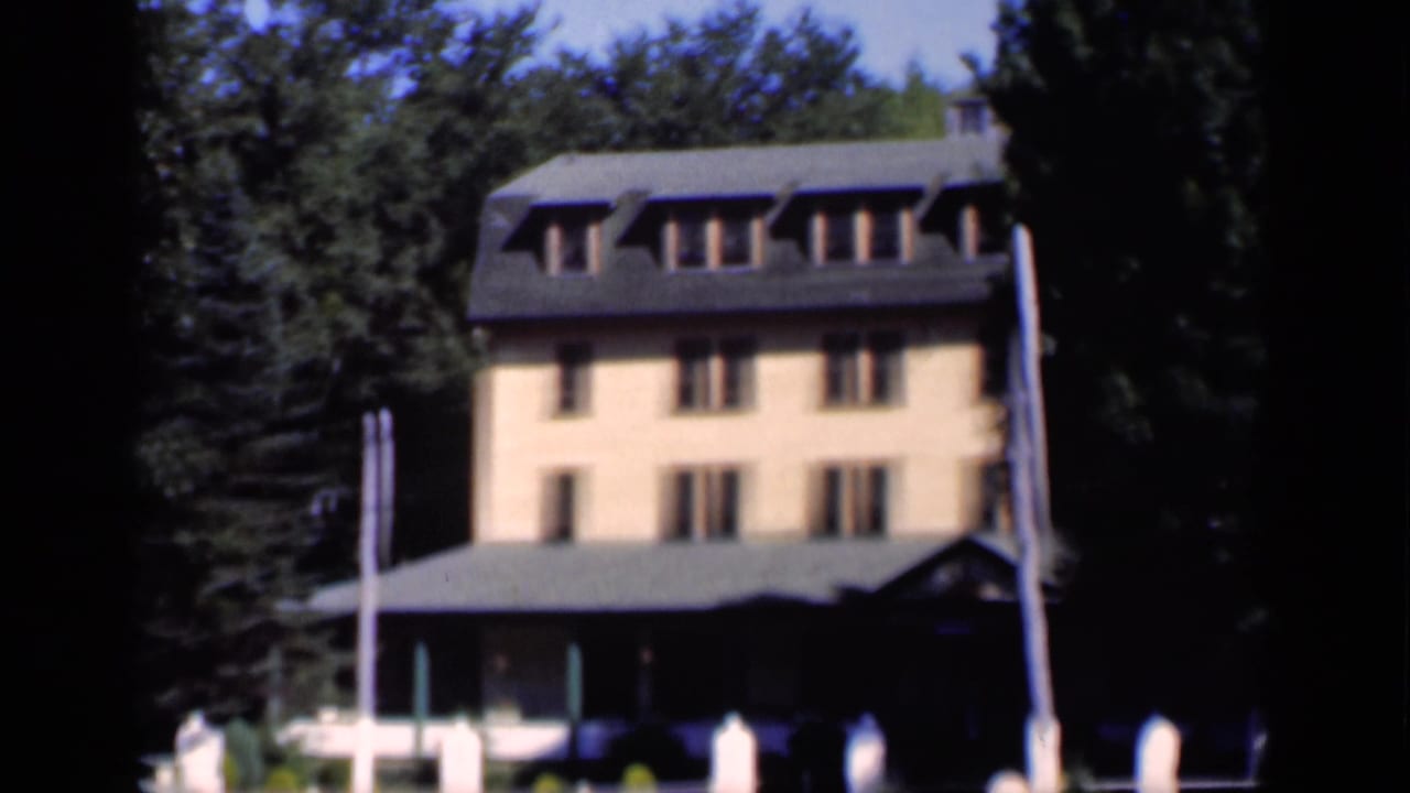 Large yellow-orange building with dormer windows partly framed by trees and a white fence.