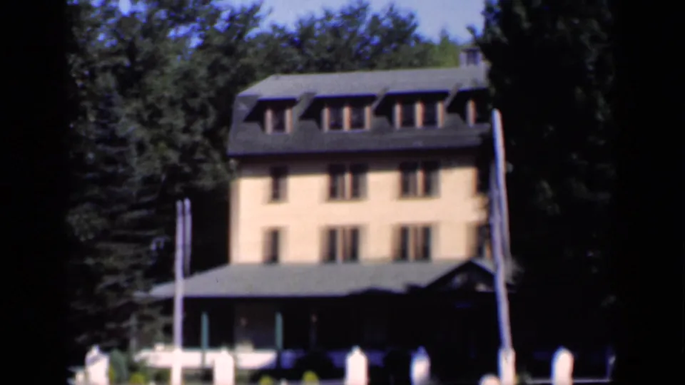 1951 New York exterior frame with a yellow-orange building, dark roof dormers, trees, lawn, and white fence elements.