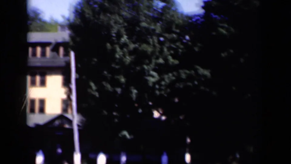 1951 New York 8mm frame showing a partial yellow-orange building, dense trees, a pole, and dark edge framing.