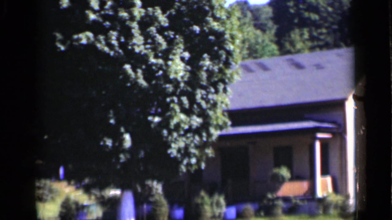 1951 New York 8mm frame showing a pale house, shaded porch area, green lawn, and large foreground tree.