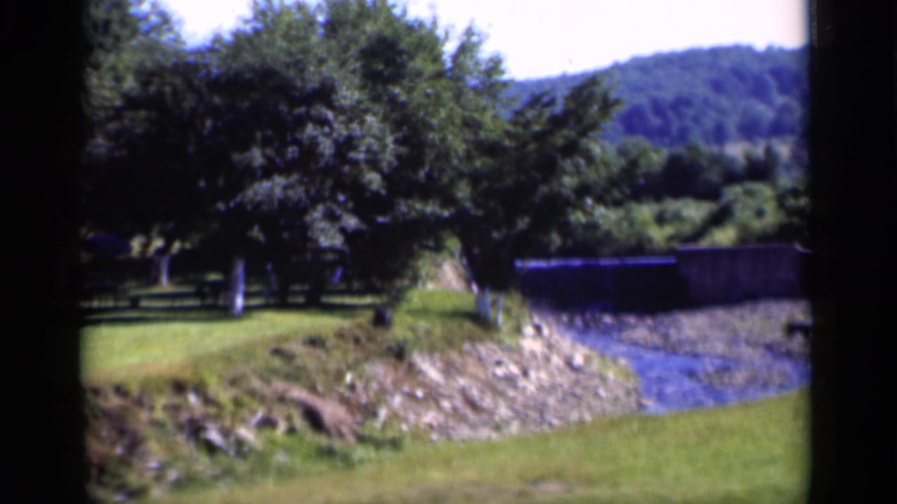 Rural lawn, trees, rocky creek bed, and wooded hills seen through dark film-frame edges.