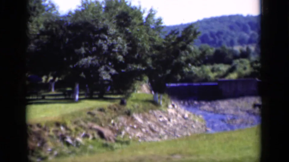 1951 New York home-movie frame showing green grass, trees, a narrow rocky creek, and wooded hills in the distance.