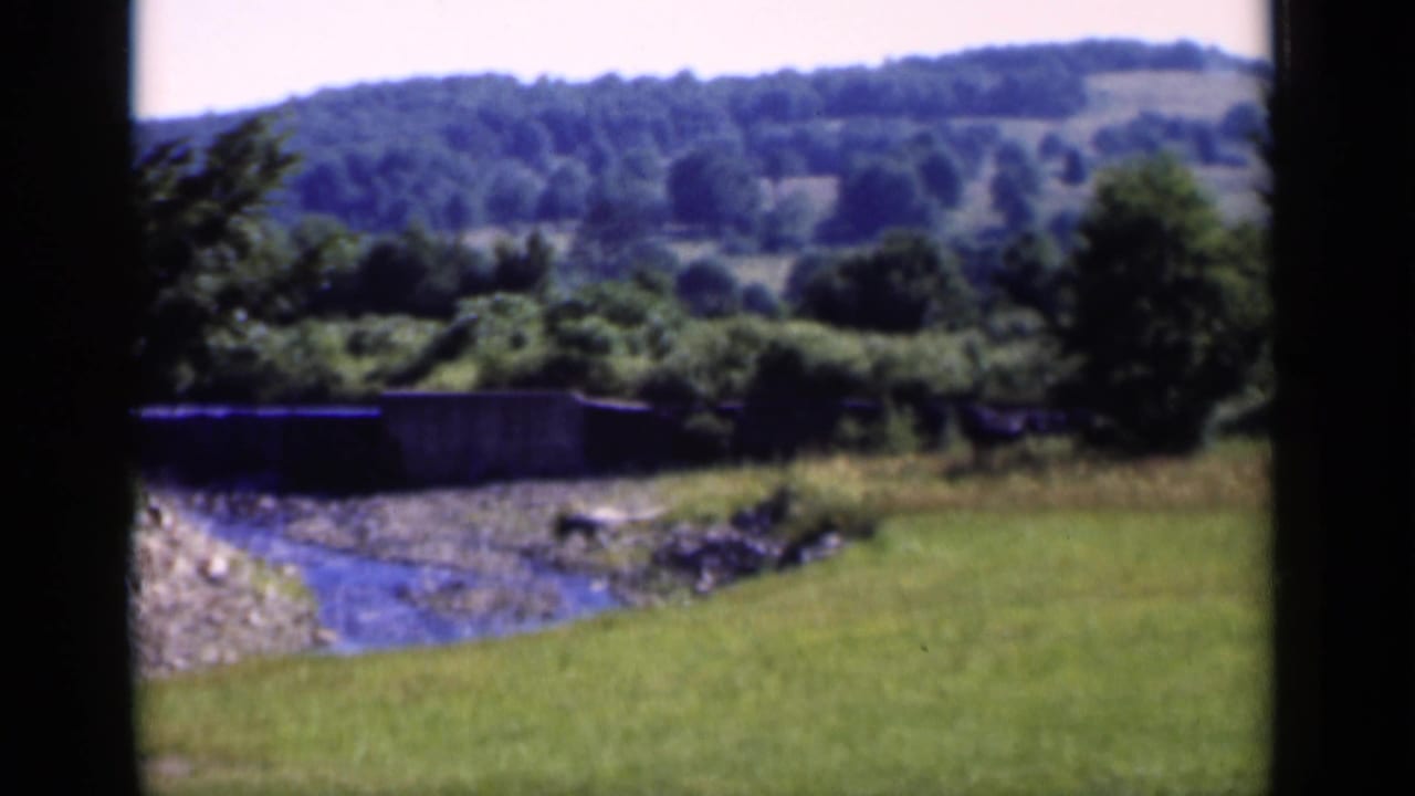 Rural New York landscape frame with a small creek, green lawn, trees, and distant wooded hills, filmed in 1951.