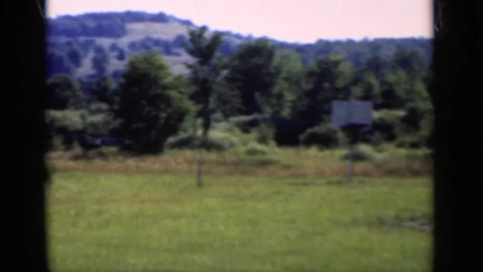 1951 New York home-movie view of a grassy rural field with trees, hills, and a basketball hoop visible near center.