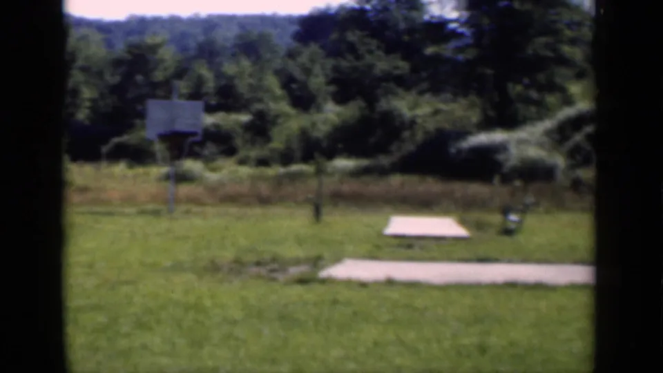 Rural New York frame from 1951 showing a yard with grass, a basketball hoop, paved slabs, trees, and wooded hillside.