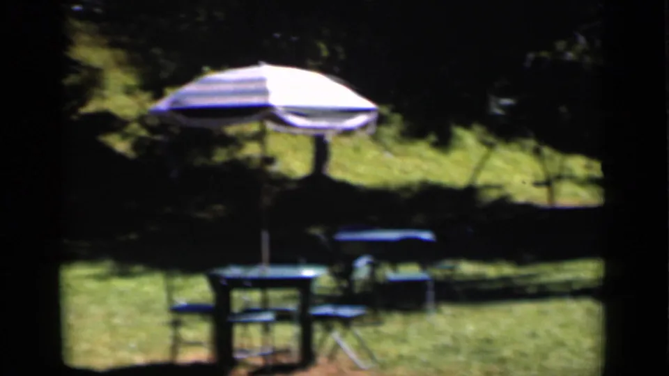 1951 New York home-movie frame showing a striped patio umbrella, folding chairs, a small table, and lawn furniture in shade.