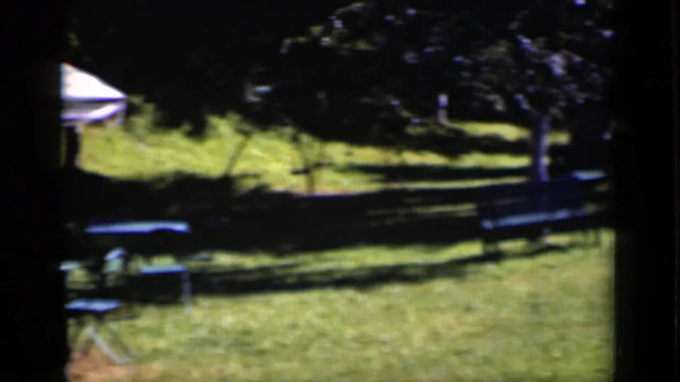 1951 New York home-movie frame of a sunlit lawn with patio umbrella, small furniture, trees, and bench seating.