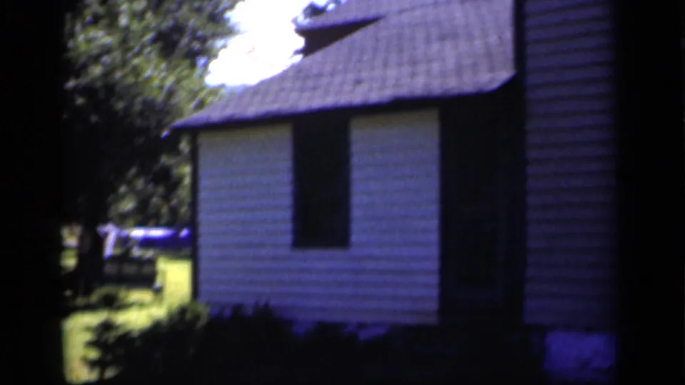 1951 New York home-movie frame showing a white-sided house exterior, dark window, roofline, shrubs, and lawn beyond.