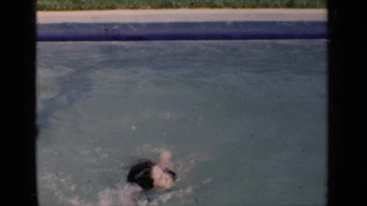 A child in a dark swimsuit swims through a backyard pool, partly turned in the water, with visible splashing and 8mm frame borders.