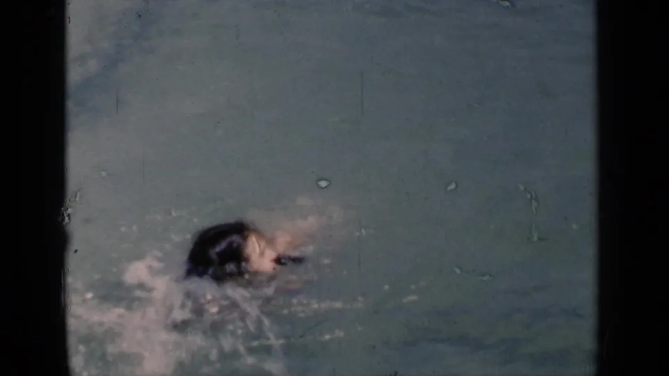 A young swimmer is partly submerged in the pool, moving across the frame in soft-focus 8mm home-movie footage.