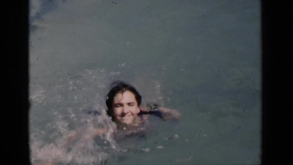 A child swims toward the camera and smiles in a 1951 New York backyard pool scene, with splashing water around the face.
