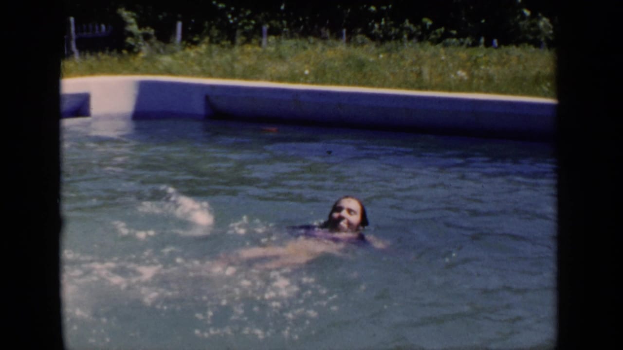 Swimmer smiling while paddling in an outdoor pool near a grassy edge.