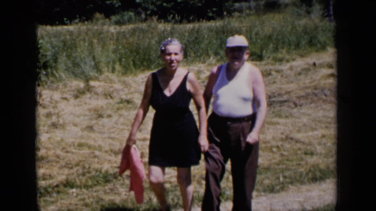 The couple continues walking toward the camera in a sunlit New York field, with the woman carrying a pink cloth and the man wearing a light cap.