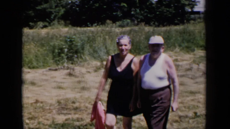 A closer home-movie frame shows the older couple smiling as they walk through dry grass, with trees and tall vegetation behind them.