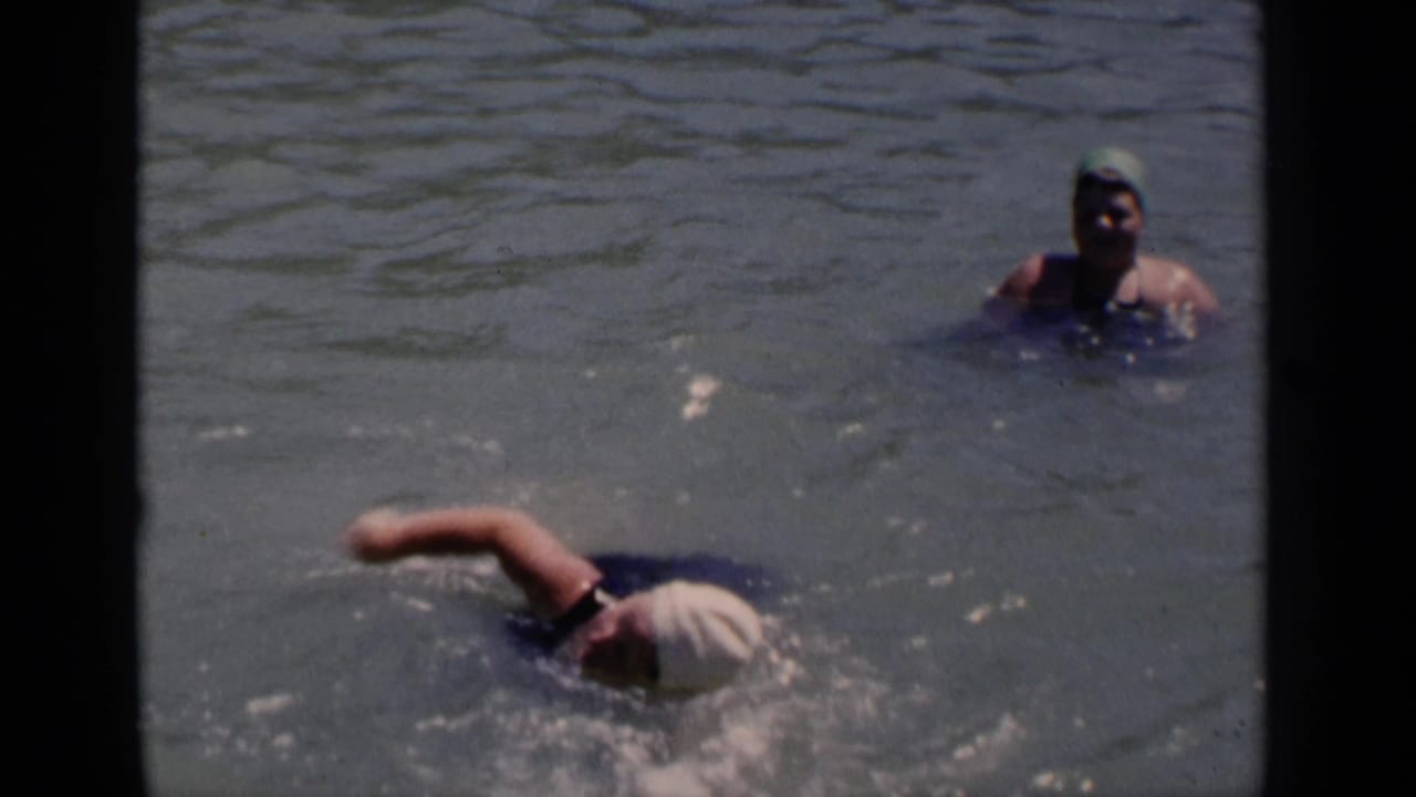 1951 New York home-movie frame of a capped swimmer moving through sunlit water while another swimmer remains upright behind her.