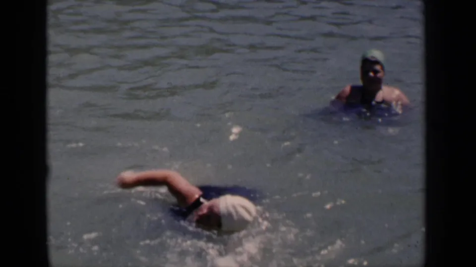 1951 New York home-movie frame of a capped swimmer moving through sunlit water while another swimmer remains upright behind her.