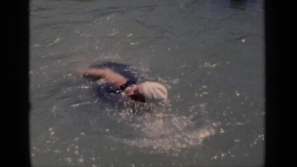 1951 New York home-movie frame showing the swimmer in a white cap splashing through open water during a freestyle stroke.