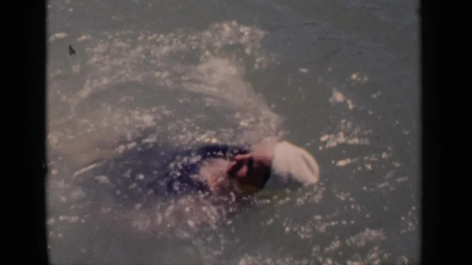 1951 New York home-movie frame showing a woman swimmer partly turned in open water, with her white cap and arm visible through spray.