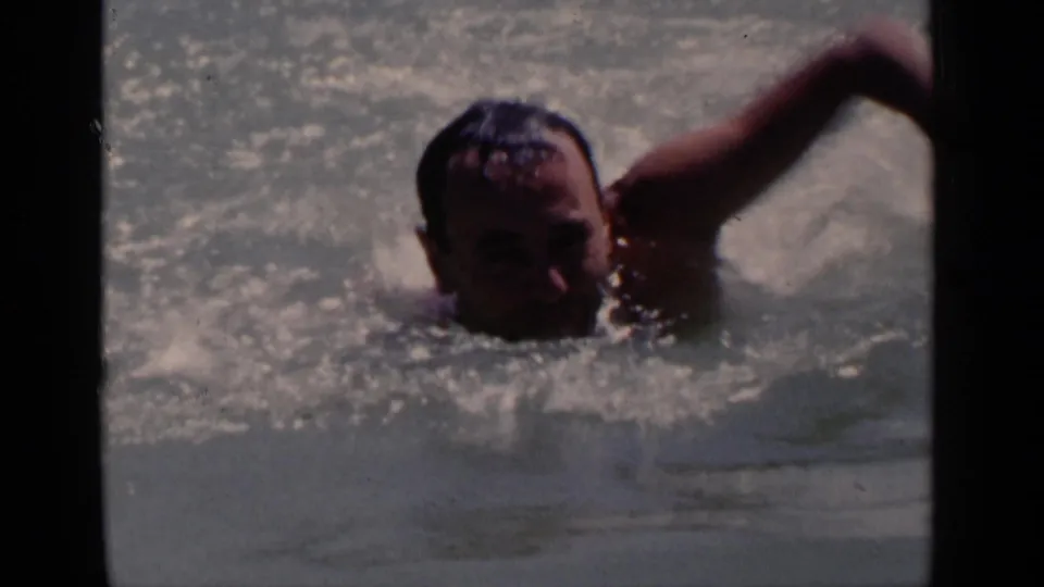 Close view of the swimmer in a vintage 8mm pool scene, face partly visible as one arm pulls through the water.