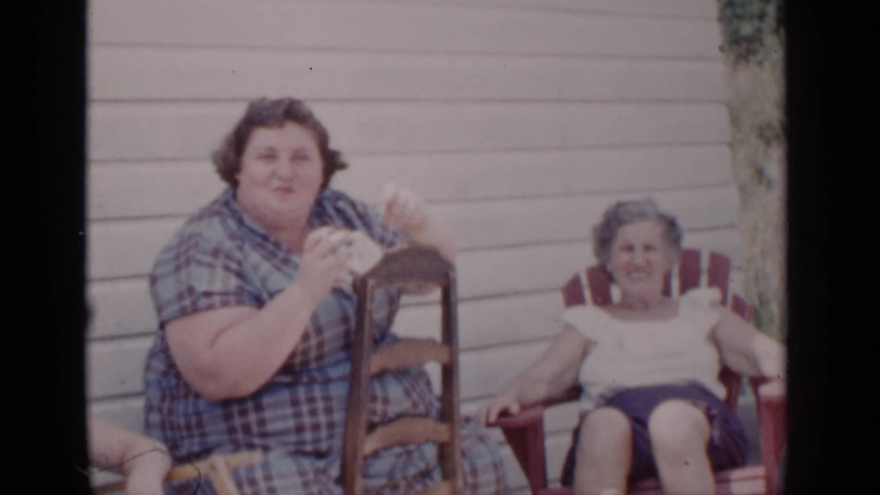 Two adult women sit outside near porch siding while one holds a small object.