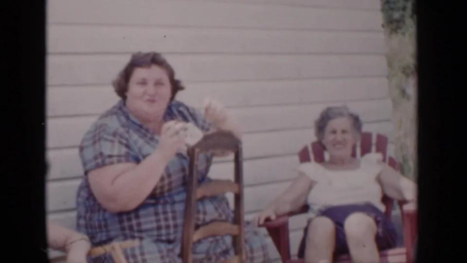 In 1951 New York home-movie footage, two adult women sit outdoors as one in a plaid dress gestures with a small object.