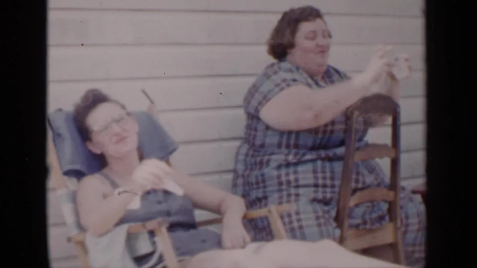 An adult woman in glasses reclines in an outdoor chair as a woman in a blue plaid dress sits nearby in this 1951 New York home-movie frame.