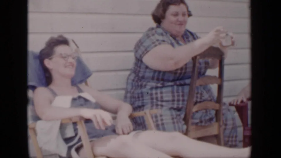 Two adult women sit outdoors in 1951 New York footage, one smiling in a striped dress while another in plaid gestures beside a chair.