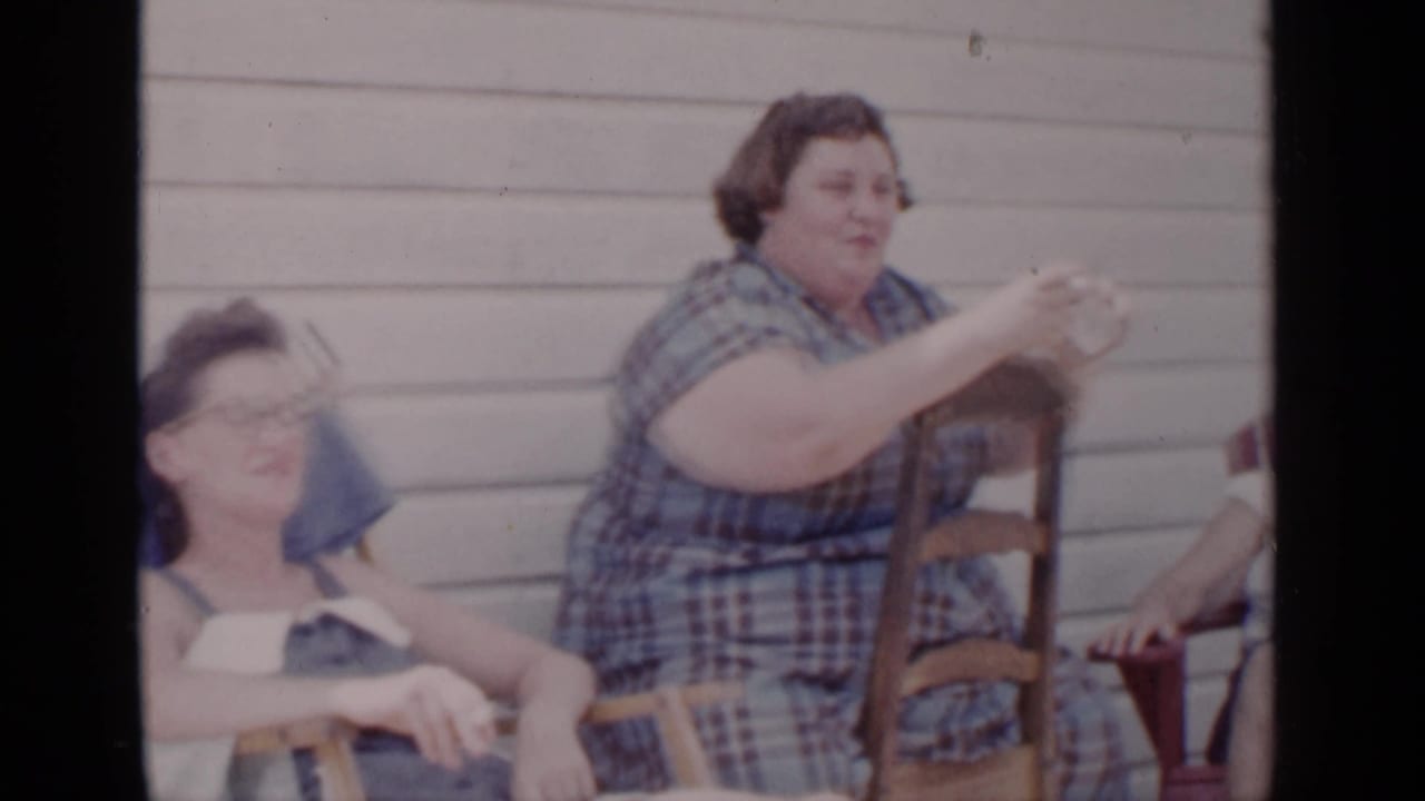 In 1951 New York home-movie footage, women sit outdoors against clapboard siding while a woman in a blue plaid dress gestures during conversation.