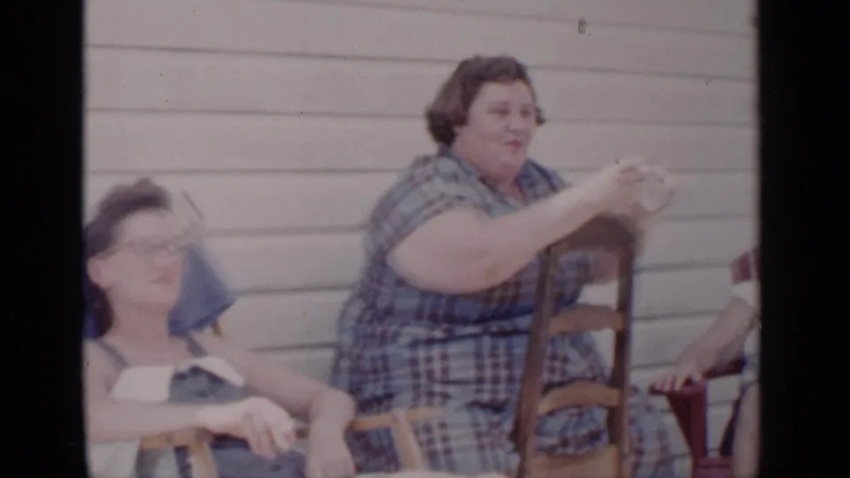 In 1951 New York home-movie footage, women sit outdoors against clapboard siding while a woman in a blue plaid dress gestures during conversation.