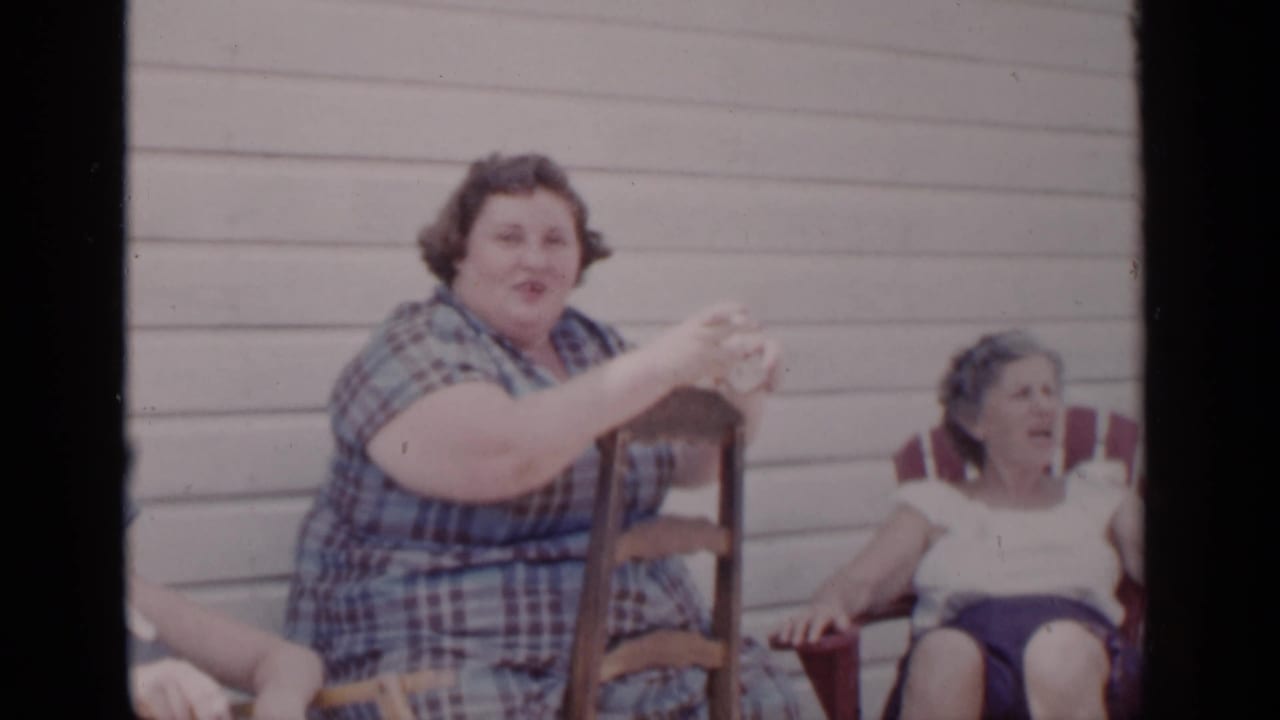 A woman in a plaid dress gestures while another woman sits in a red chair beside her.