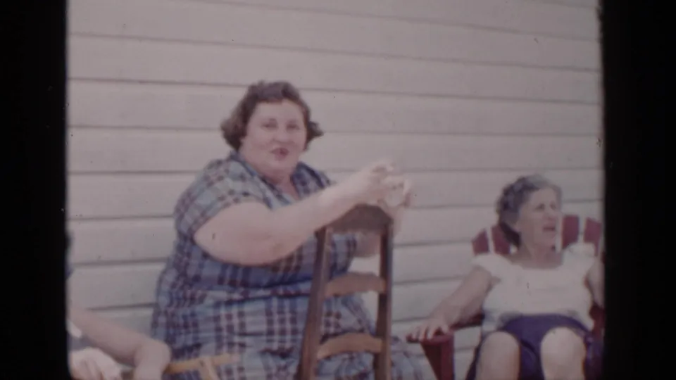 In 1951 New York home-movie footage, a woman in a plaid dress gestures while a seated woman in a light top and dark shorts looks on from a red chair.